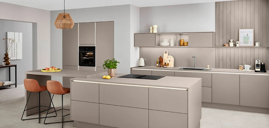 Line One 01 Grifflose Küche in Taupe mit Kochinsel, angesetzter Theke mit zwei Barhockern und hinterleuchteter waagerechter Griffspur