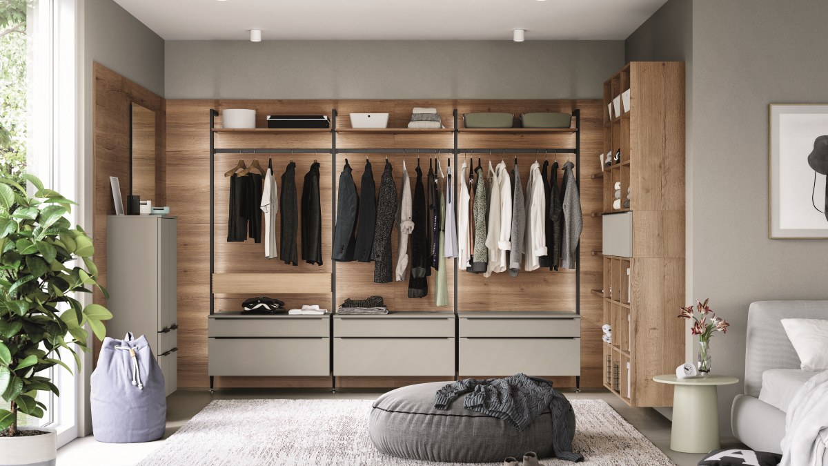 Offene Kleiderschrankwand mit Holzrückwand, Kleiderstangen, grauen Schubkastenelementen und Regalturm neben einem Bett